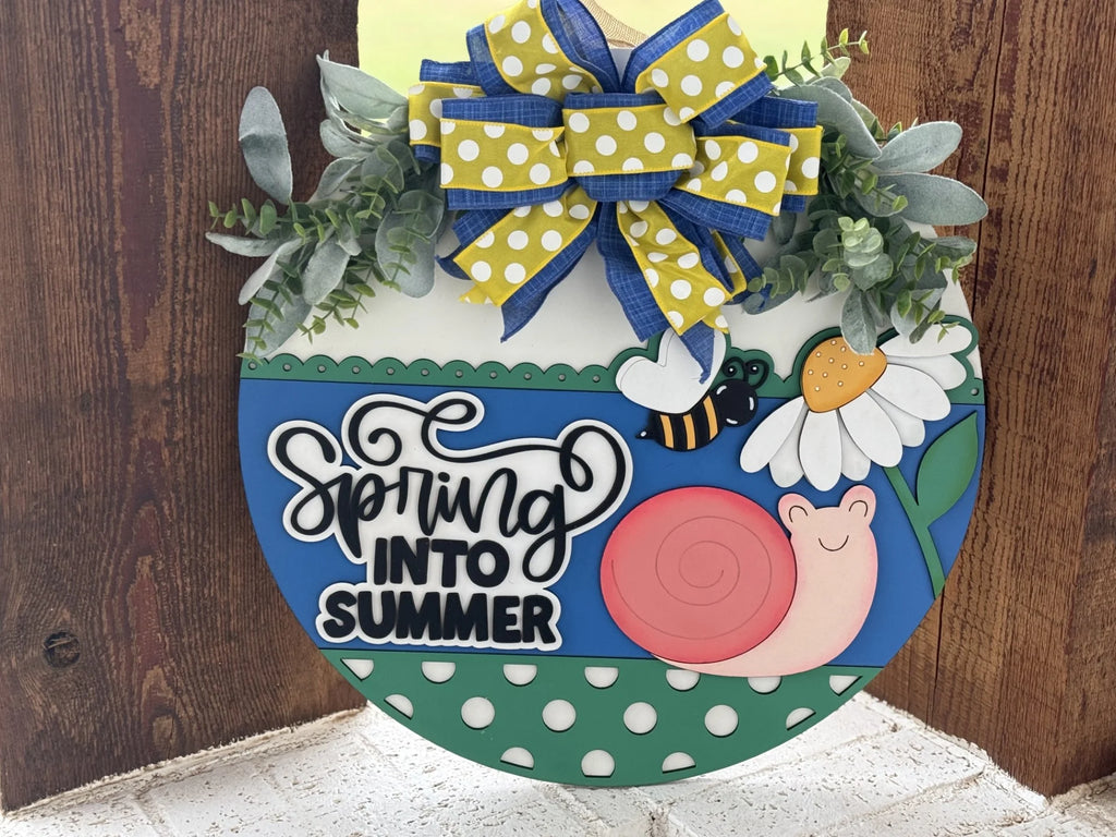 A colorful round sign with the text Spring into Summer features a cartoon pink snail, a bee, and a daisy. The top is adorned with a bow of blue and yellow polka-dotted ribbons, surrounded by green leaves. The bottom has a green polka-dotted pattern.