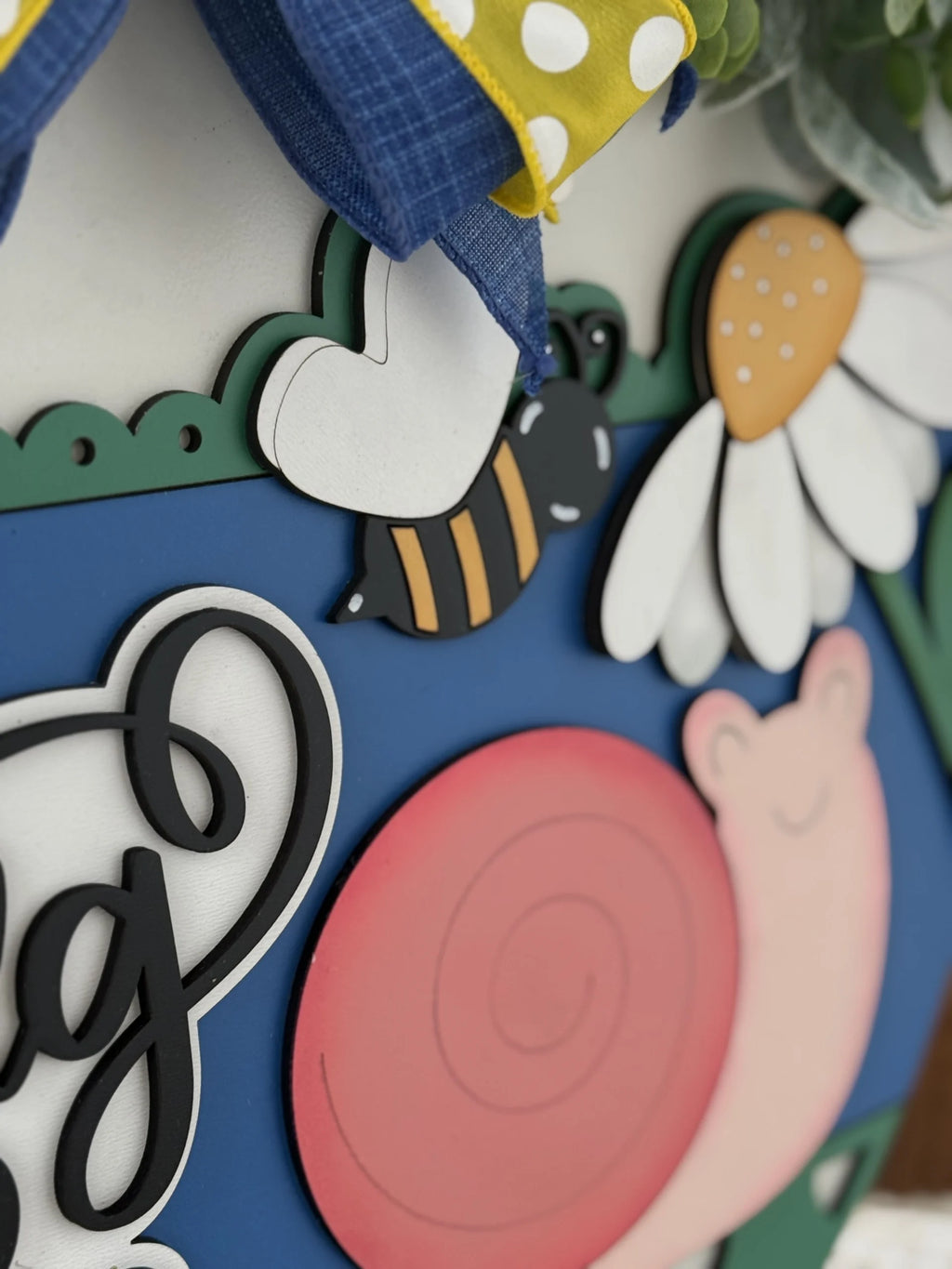 Close-up of a colorful decorative wall hanging featuring a snail with a pink shell, a bee, and a white daisy with a yellow center on a blue background. Theres a scalloped green edge above, and a blue and yellow polka-dotted ribbon on top.
