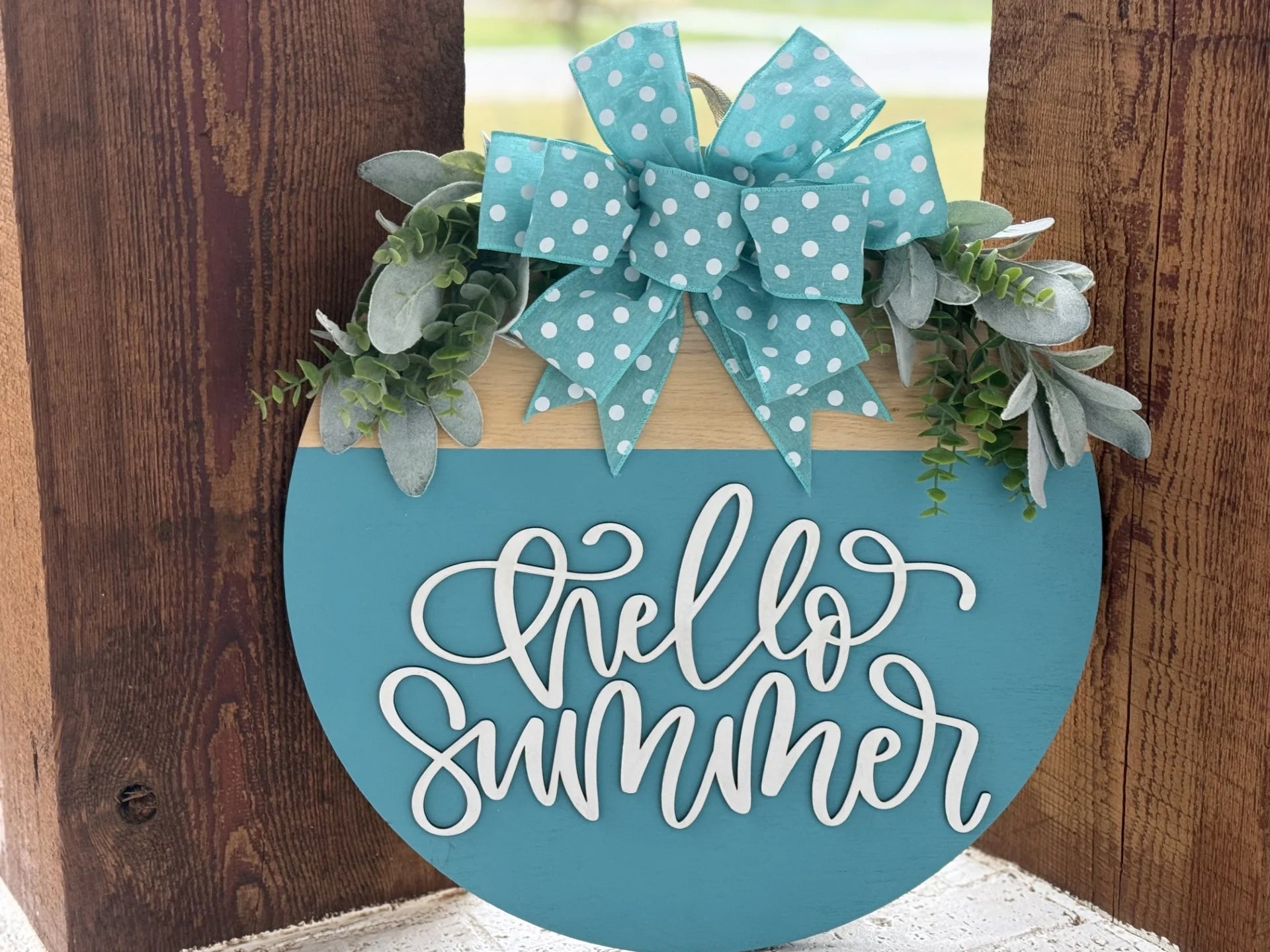 A round teal sign with the white cursive phrase hello summer is adorned with a teal polka-dotted bow at the top. Greenery accents surround the bow. The sign is propped up against a wooden surface with a blurred outdoor background.