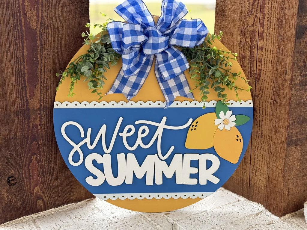 A round sign with a yellow and blue color scheme hangs on a wooden surface. It features the words Sweet Summer in white script. Above, theres a blue and white gingham bow, and beside it, small sprigs of greenery and lemon illustrations enhance the design.