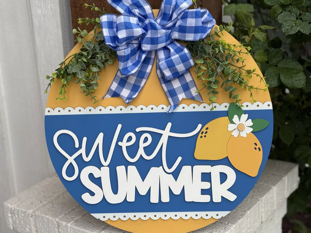 A decorative round sign reads Sweet Summer in white text. It features painted yellow lemons with a small white flower. A blue and white gingham bow adorns the top, with green foliage on either side. The sign has a yellow and blue color scheme and rests on a white surface.