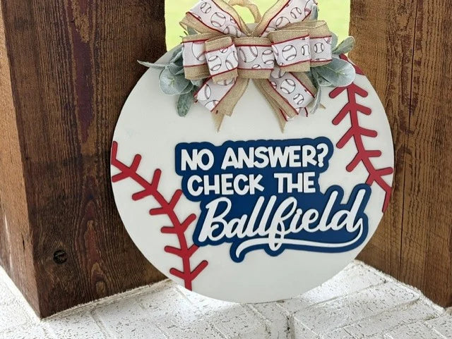 Round sign resembling a baseball hangs on a wooden surface. It features the text, No answer? Check the Ballfield in blue and white lettering. A bow with baseball-patterned ribbon and green leaves is at the top. Red stitching lines on the sign mimic a baseballs design.