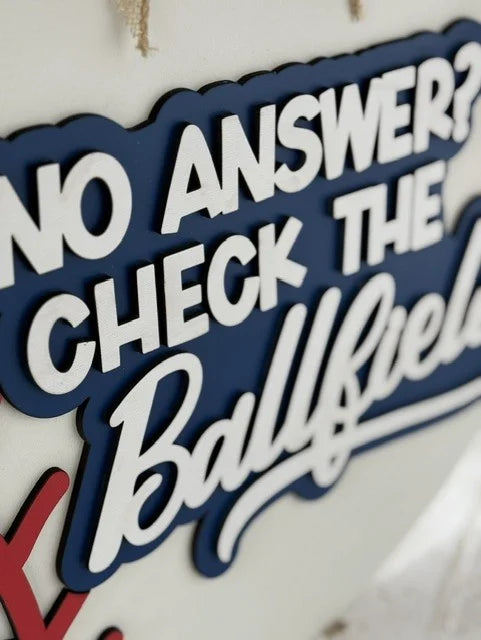 Close-up of a decorative sign with layered text. Bold white letters on a blue background read, NO ANSWER? CHECK THE Ballfield. The word Ballfield is in cursive. The sign has a playful and bold style, partially visible against a light-colored wall.