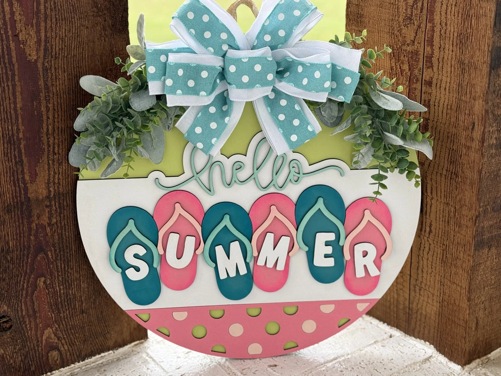 A decorative summer sign features a wooden half-circle design. It has flip-flops spelling SUMMER and hello in cursive. Greenery and a polka-dotted teal bow adorn the top. The colors are vibrant, including pink, blue, green, and white, set against a wooden background.