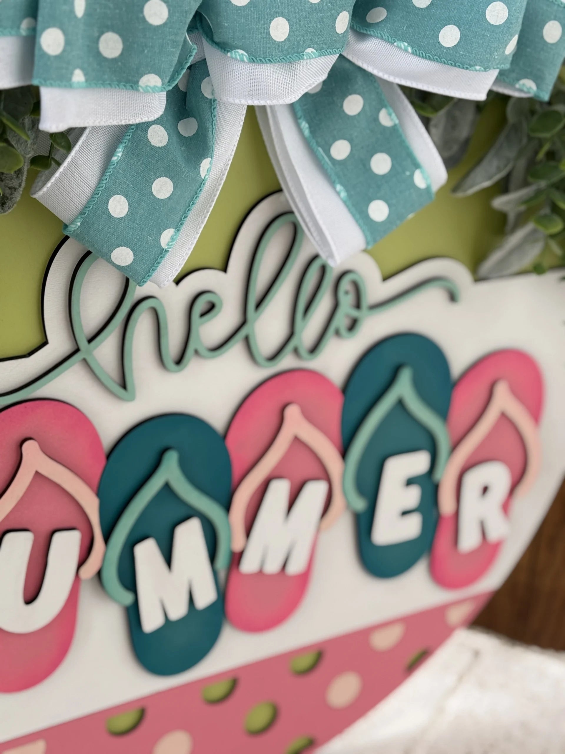 Close-up of a decorative sign featuring the text hello SUMMER in playful fonts. Summer is spelled with letters placed on colorful flip-flop shapes. A large, decorative bow with teal and white polka dots is above the sign, and greenery peeks out from behind.