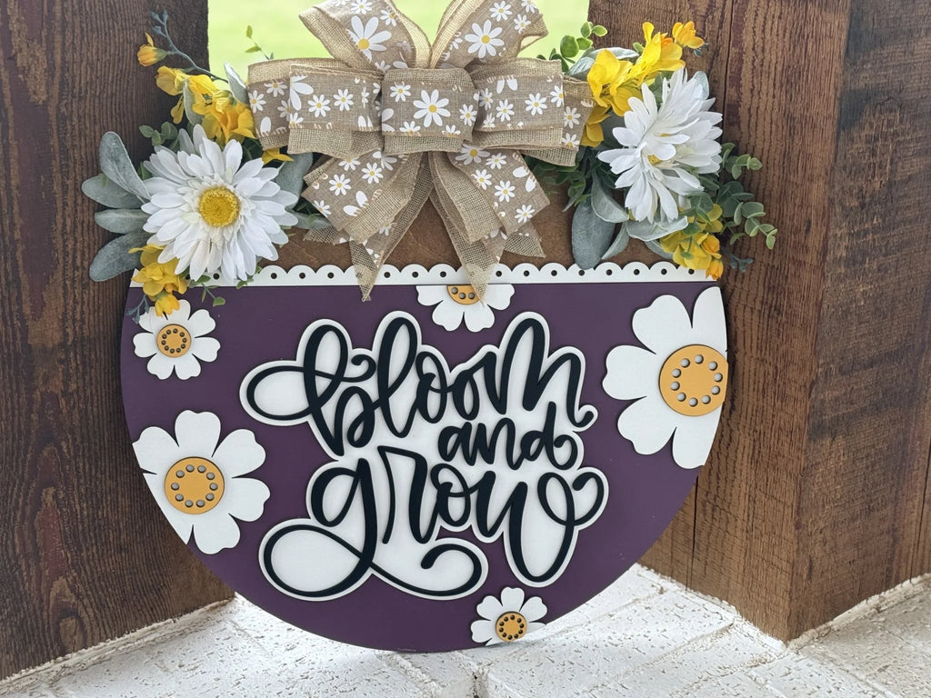 A circular sign features the phrase Bloom and Grow in stylish black script on a purple background adorned with white and yellow flowers. A burlap bow with daisy patterns sits atop, surrounded by white daisies and green foliage, all set against a rustic wooden backdrop.