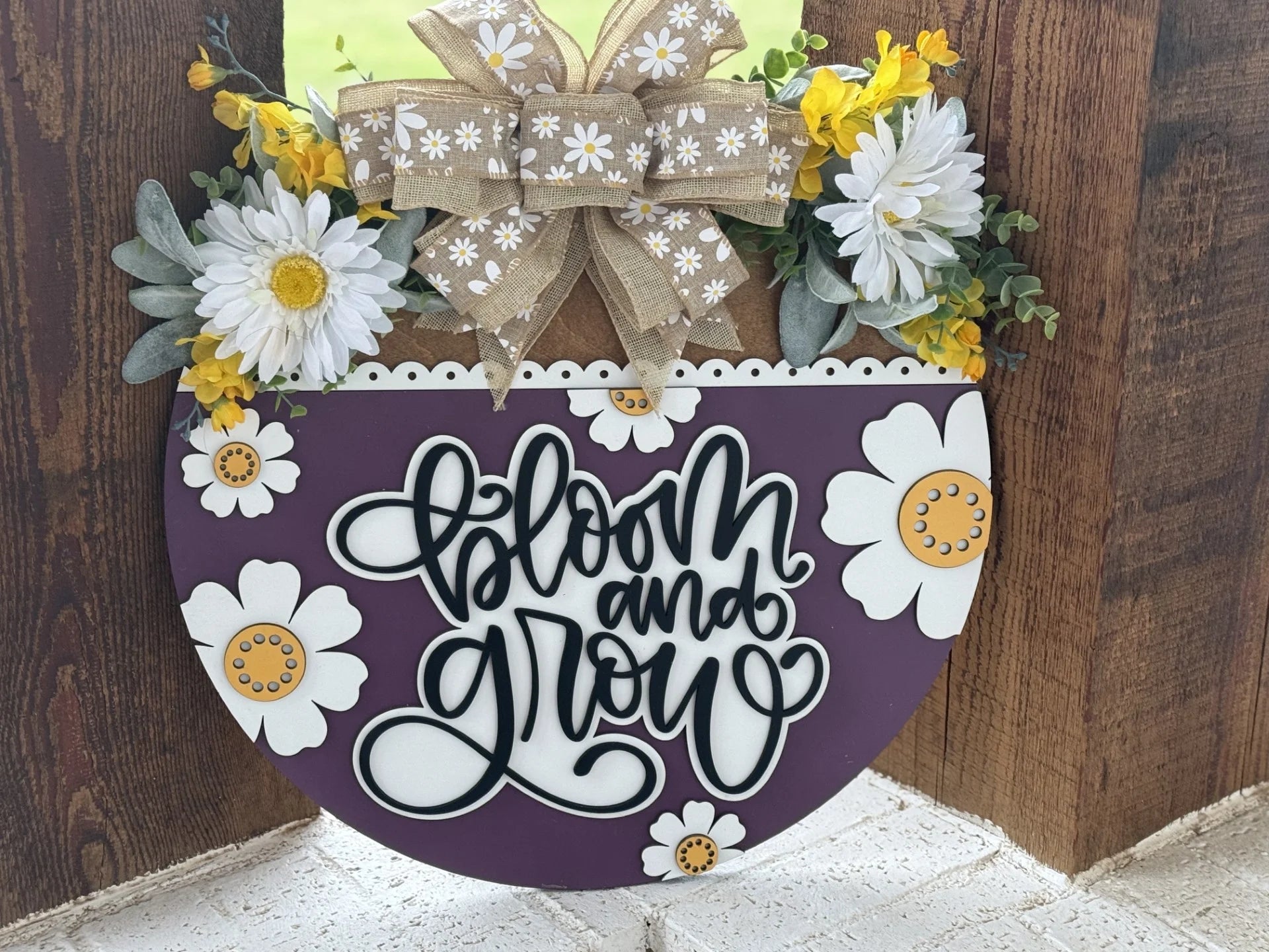 A circular sign features the phrase Bloom and Grow in stylish black script on a purple background adorned with white and yellow flowers. A burlap bow with daisy patterns sits atop, surrounded by white daisies and green foliage, all set against a rustic wooden backdrop.
