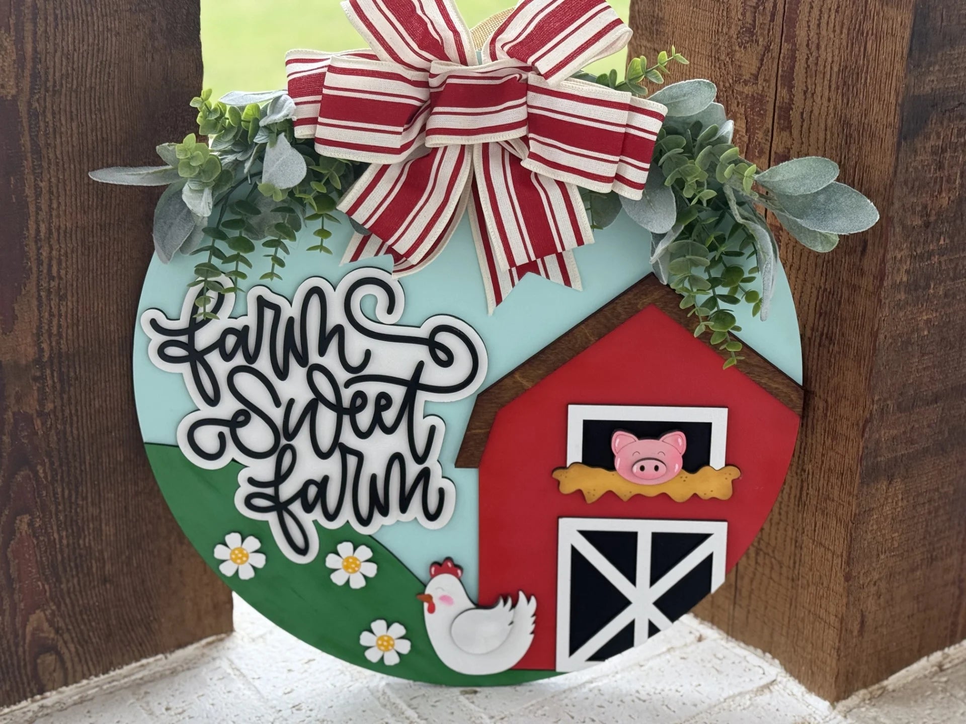 Round wooden sign with Farm Sweet Farm in cursive black letters. It features a red barn with a pig, a chicken, and daisies. Eucalyptus leaves and a large red and white striped bow adorn the top. The background includes blue sky and green grass.