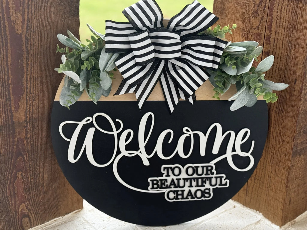 A round black sign with the text Welcome to our beautiful chaos in white script and print. The sign is adorned with green foliage and a large black-and-white striped bow at the top. It hangs on a wooden surface, with a hint of green grass visible in the background.