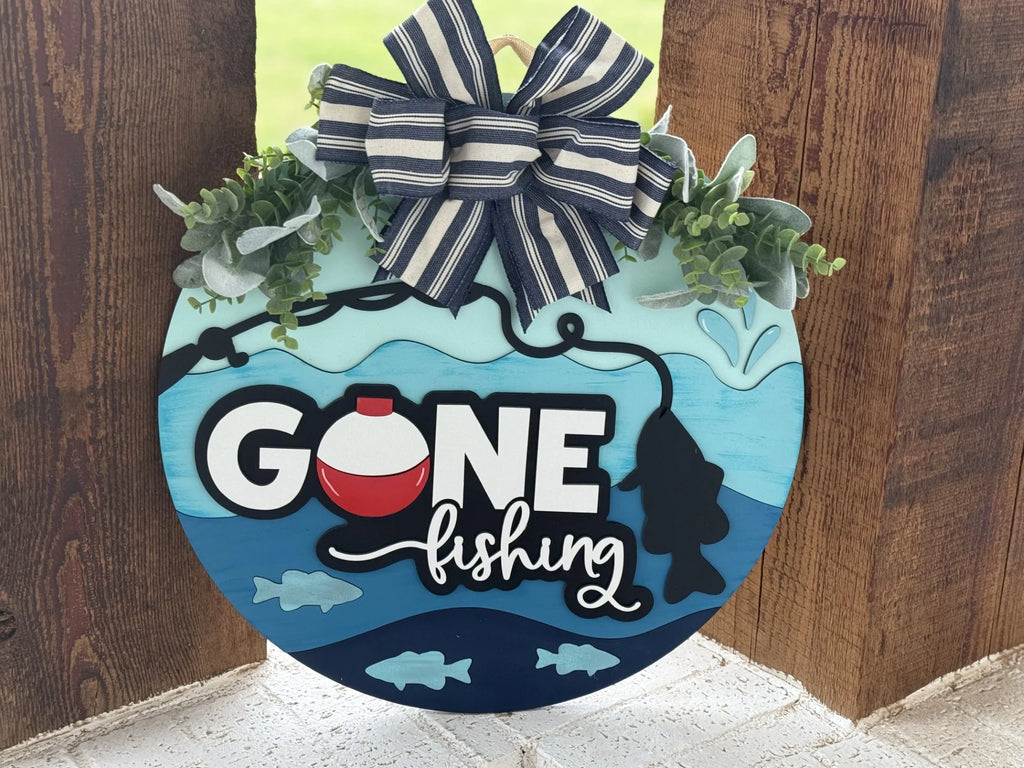 A round sign with Gone fishing written on it in bold letters. The o is a fishing bobber. The sign features a fishing rod and silhouette of a fish. Its accented with green leaves and a large blue and white striped bow on top, set against a wooden background.