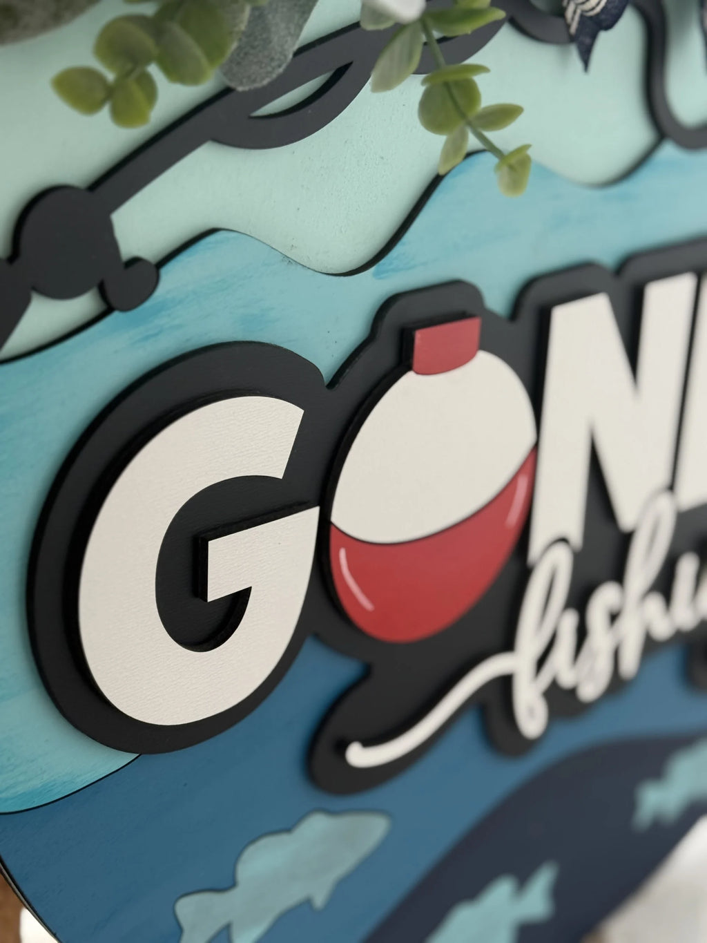 Close-up of a decorative sign featuring the words Gone fishing. The letters are bold, with a red and white fishing bobber replacing the letter o. The background is painted in shades of blue and green, with small artificial leaves adorning the top edge.