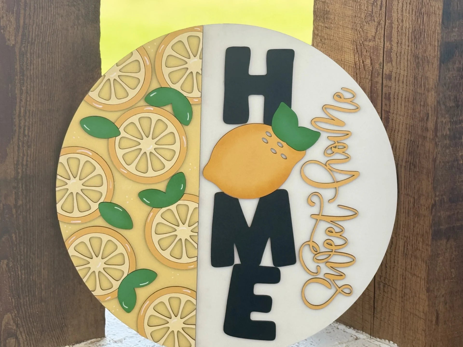 Round sign with a wooden texture background, divided in half. Left side features sliced lemons and green leaves. Right side spells HOME vertically in black, with a yellow lemon representing the O. Scripted text sweet home in yellow overlays the letters.