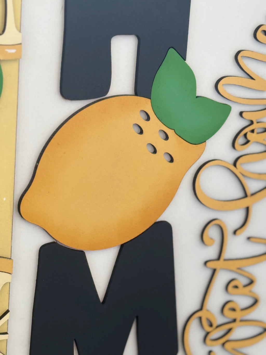 Close-up of decorative wall art featuring a stylized lemon with green leaves in the center. The lemon sits over black, blocky letters, with cursive golden text partially visible on the right. The background is a neutral off-white.