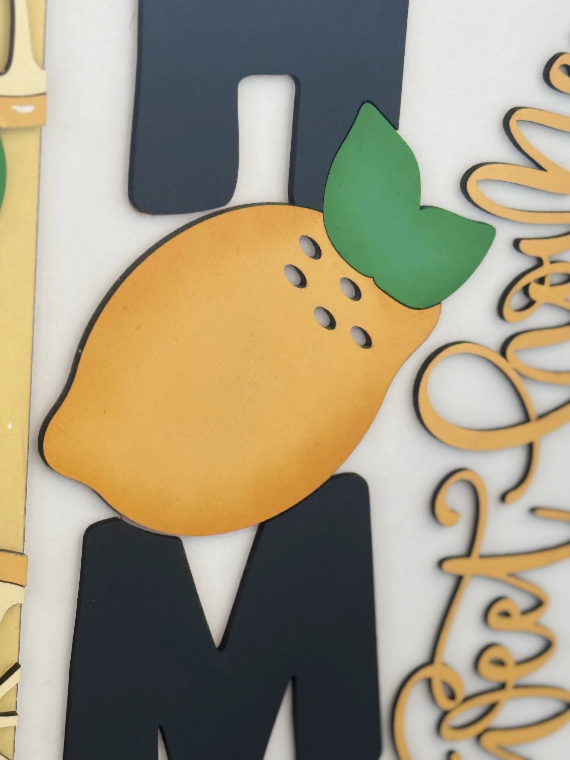 Close-up of decorative wall art featuring a stylized lemon with green leaves in the center. The lemon sits over black, blocky letters, with cursive golden text partially visible on the right. The background is a neutral off-white.