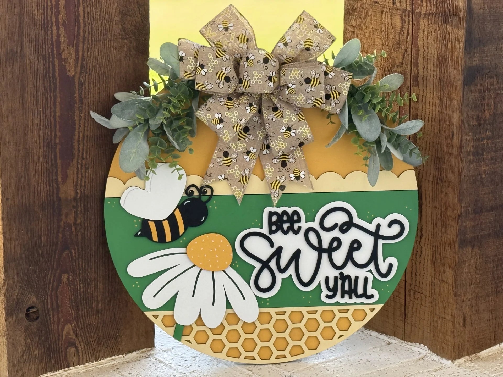 Round sign with carved text Bee Sweet Yall and a daisy motif, featuring a bee and hexagon pattern. Topped with a bow decorated with bees, greenery, and honeycomb accents on a wooden backdrop, conveying a cheerful, rustic theme.