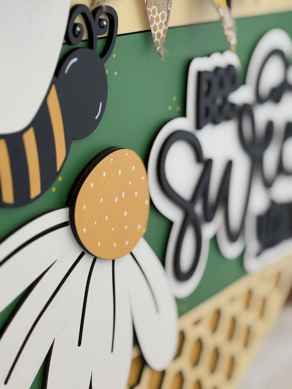 Close-up of a decorative sign featuring a stylized bee with black and orange stripes on a green background. The text Bee Sweet is partially visible. A daisy with white petals and a yellow center is in the foreground. The design includes textured and layered elements.