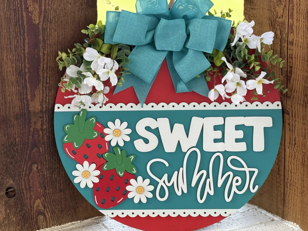 A round, colorful wooden sign with a teal and red background reads “Sweet Summer” in bold white letters. It is decorated with strawberries, white flowers, a large teal bow, and green leaves, and is hanging against a wood-paneled wall.