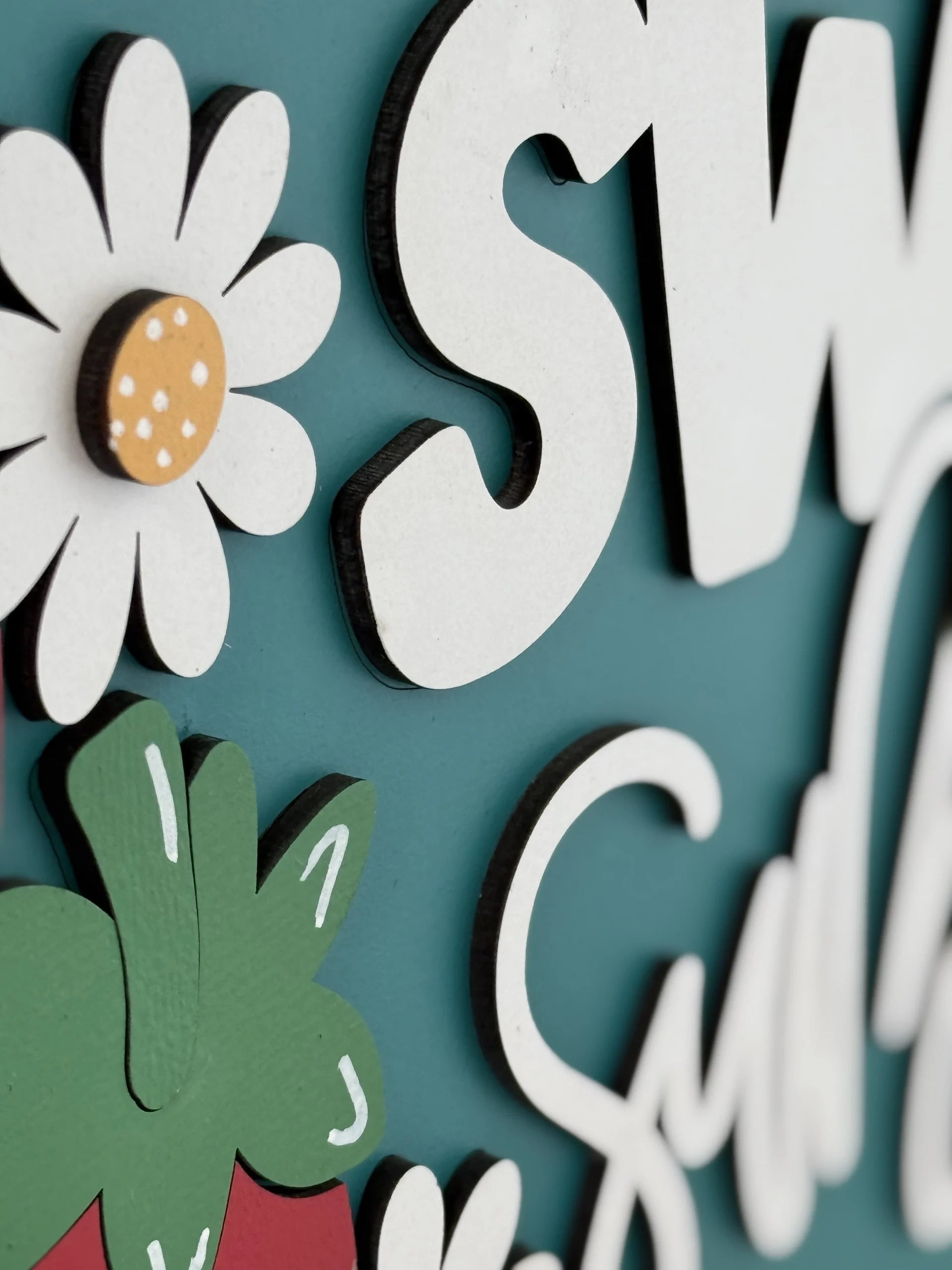 A close-up of a decorative sign with white, raised letters against a teal background. The sign features a white daisy with a yellow center and a red strawberry with green leaves, both with a playful, hand-painted style.