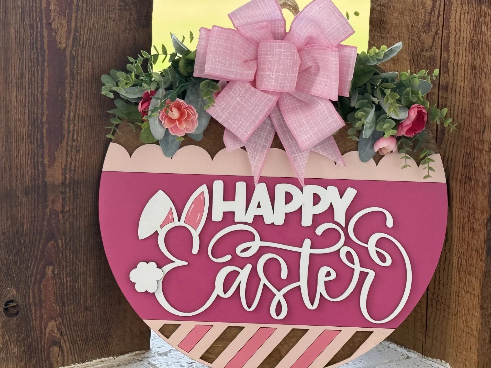 A pink egg-shaped wooden sign decorated with faux greenery, pink flowers, and a large pink bow. The sign reads Happy Easter in white script, with a bunny ear and a small bunny tail accent on the word Easter.