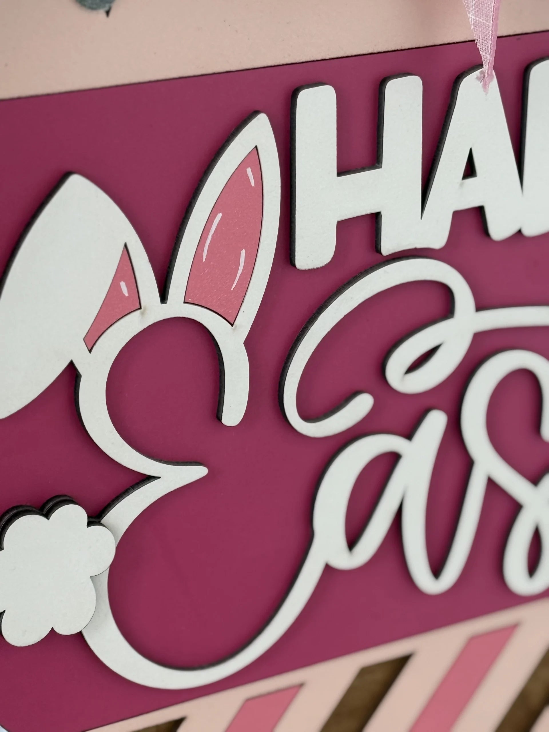 Close-up of a decorative sign with white and pink letters spelling Happy Easter. The E is styled as a bunny head, featuring bunny ears and a fluffy tail. The background is a bold magenta color with some light pink and beige accents.