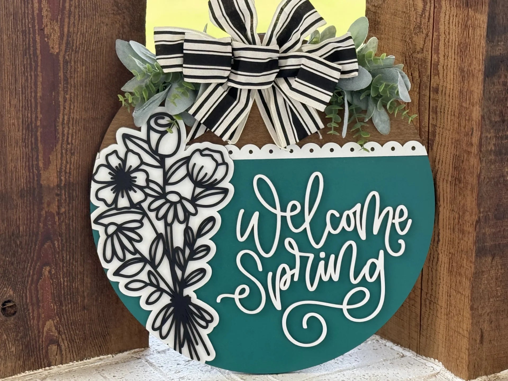 A round wooden sign with teal and white paint hangs on a wall. It features black and white floral designs on the left, the words Welcome Spring in elegant script, a black and white striped bow, and faux greenery at the top.
