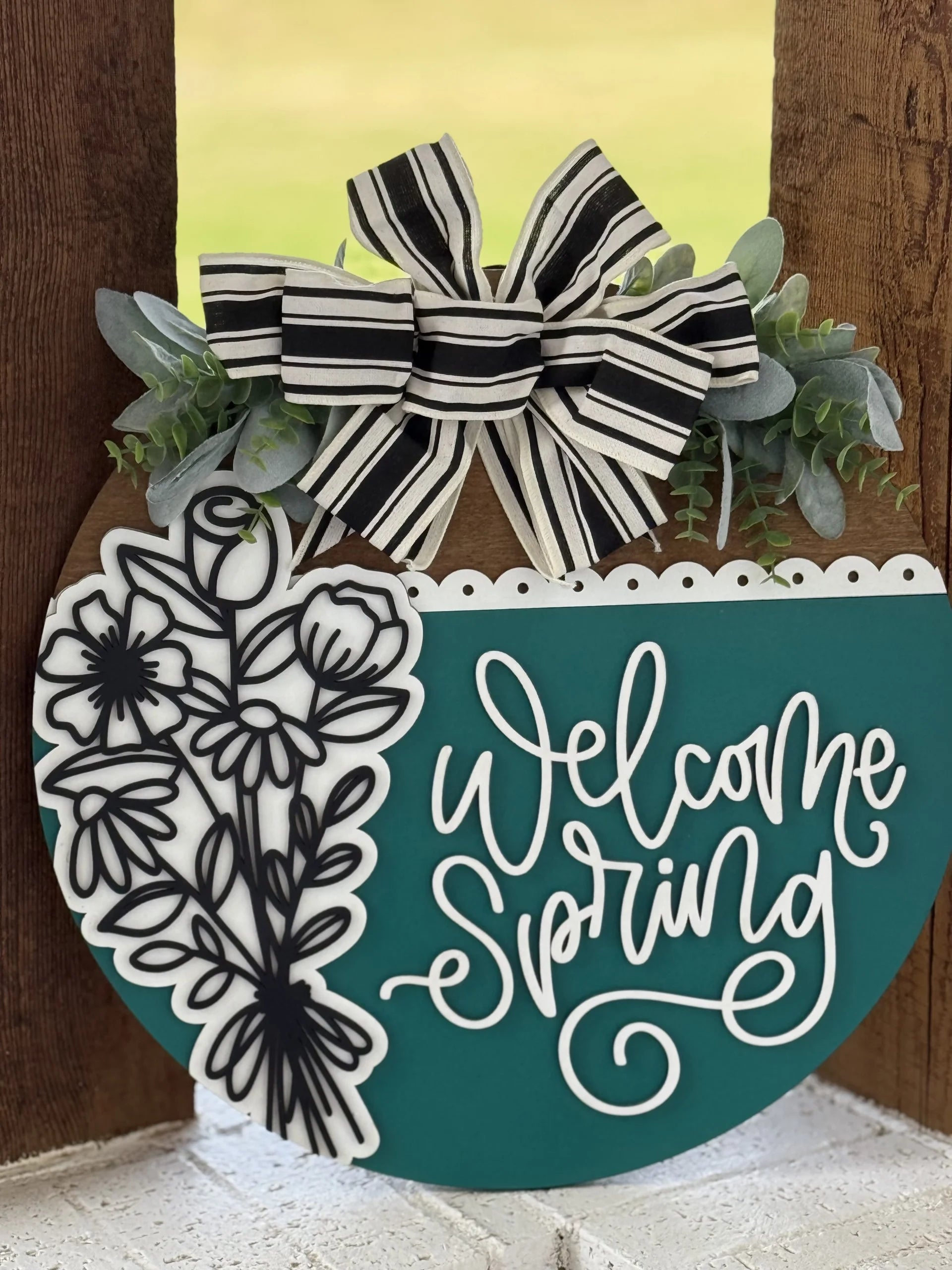 A round wooden sign with a teal lower half reads “Welcome Spring” in white script. The top is decorated with black-and-white striped ribbon, faux greenery, and white and black floral designs. The sign hangs outdoors against a wooden surface.