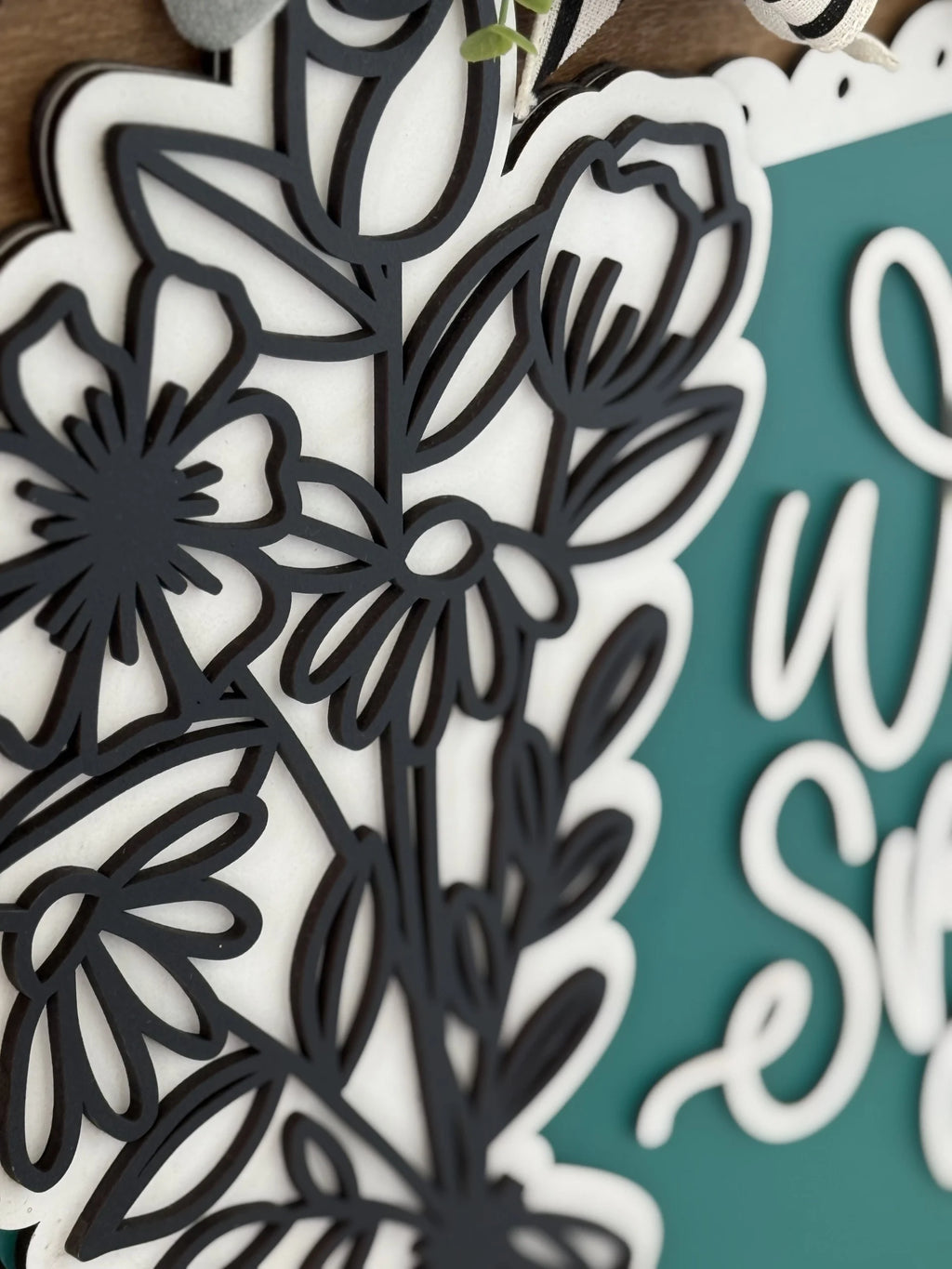 Close-up of a decorative floral design in black, layered over a white background. The flowers and leaves are detailed and appear in silhouette. To the right, there is a partial view of teal-colored signage with cursive white text.