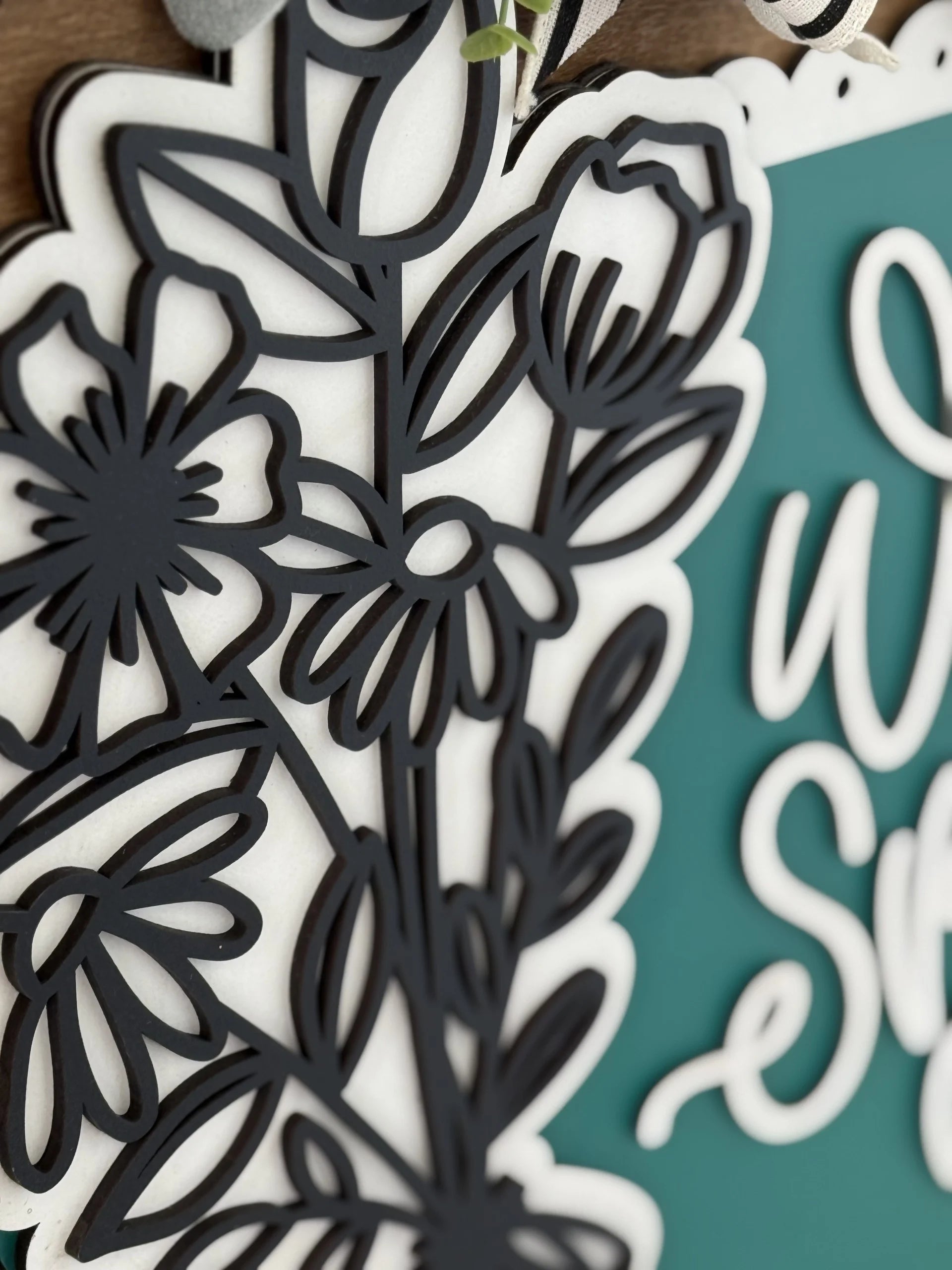 Close-up of a decorative floral design in black, layered over a white background. The flowers and leaves are detailed and appear in silhouette. To the right, there is a partial view of teal-colored signage with cursive white text.
