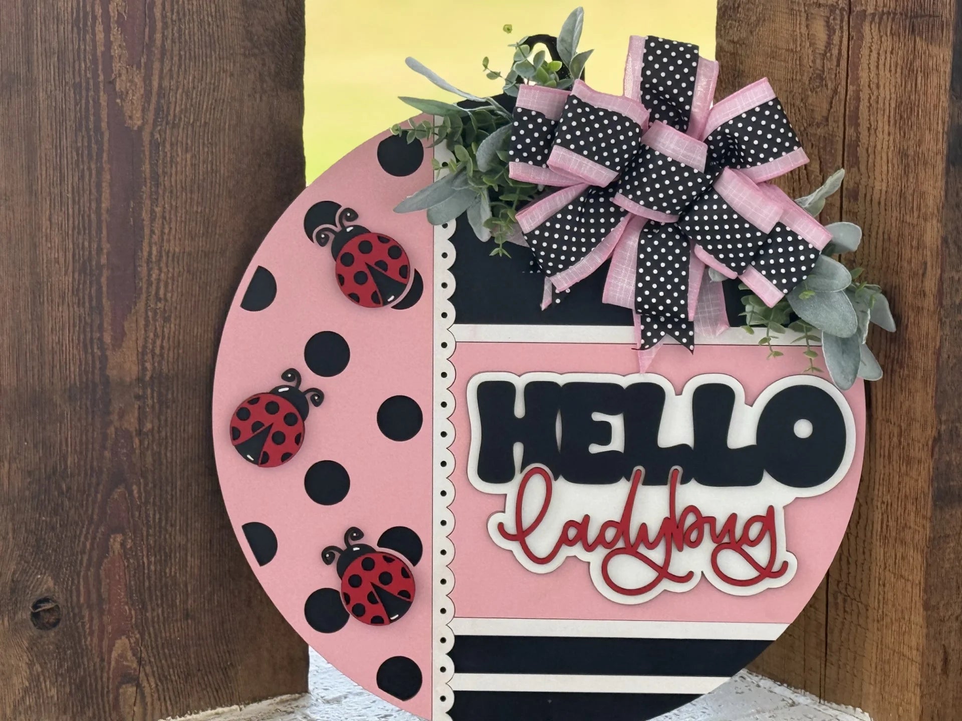 A round wooden sign with pink, black, and white stripes and polka dots, decorated with two red ladybugs, faux greenery, a large pink and black ribbon, and the words “HELLO ladybug” in bold, playful letters. The sign leans against a wooden background.