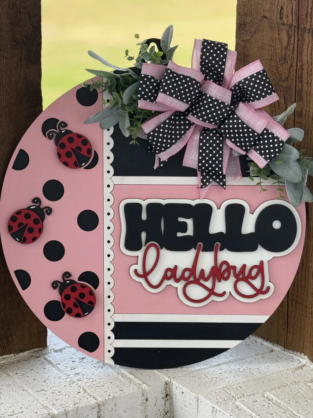 A round pink and black door sign with “HELLO ladybug” in bold letters, three red ladybug decorations, a large black, pink, and white polka-dot bow with greenery, and decorative stripes and scalloped trim on a wooden background.