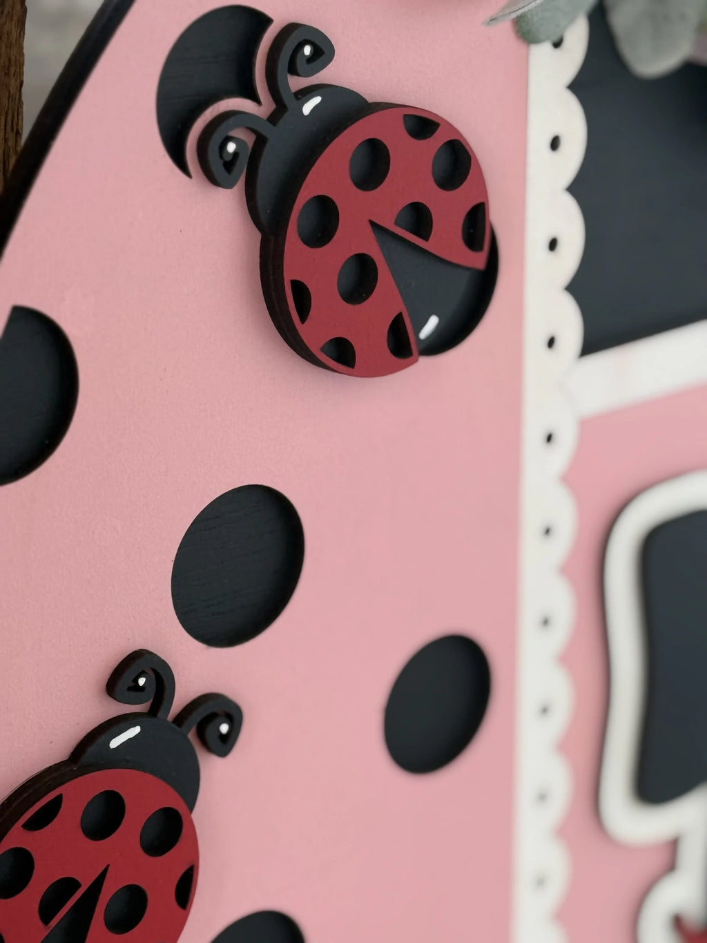 Close-up of a pink surface decorated with black polka dots and two 3D ladybug shapes. The ladybugs are red with black spots and heads, positioned near a white scalloped edge and part of a black rectangular shape.