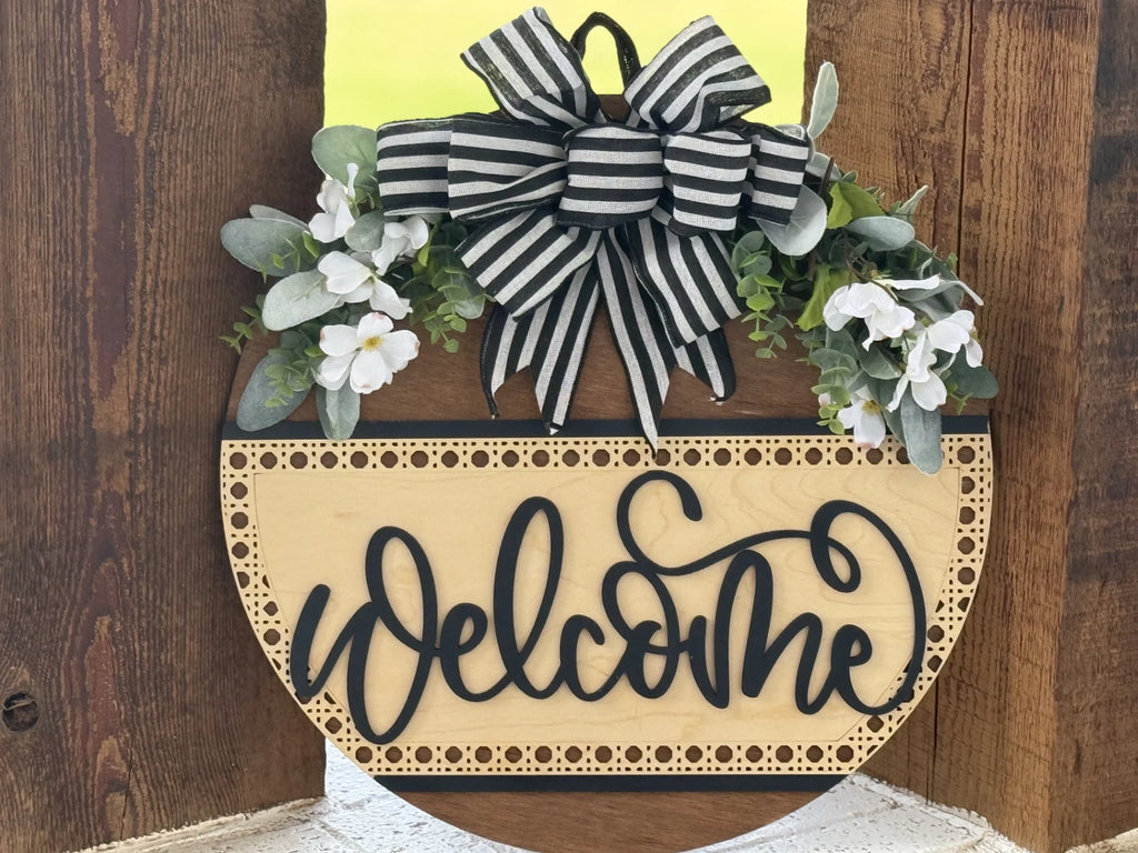 A round wooden sign with “Welcome” written in black script. The top is adorned with a black and white striped bow, green leaves, and white flowers. The sign is against a rustic wooden background.