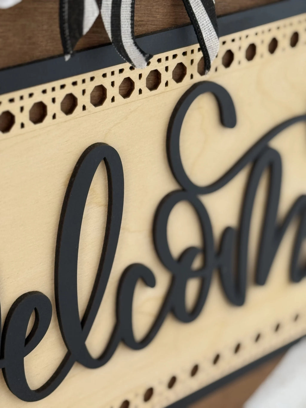 Close-up of a decorative wooden sign featuring the word Welcome in black, cursive letters. The sign has a light wood background, intricate cutout detailing along the border, and a black-and-white striped ribbon at the top.