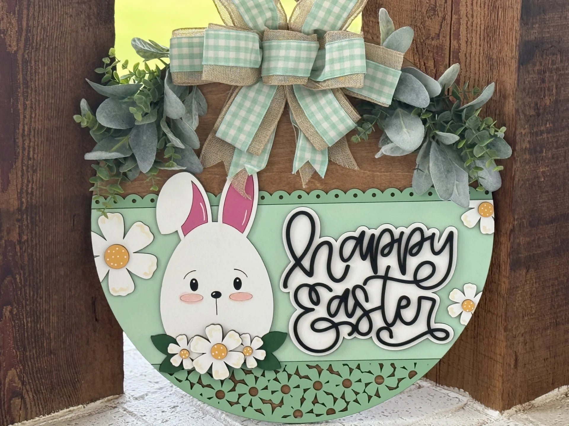 A round wooden Easter sign with a cute white bunny, daisies, green leaves, and the words “happy easter” in cursive. The sign is mint green with decorative cutouts, topped with a gingham and burlap bow, and propped against a wooden background.