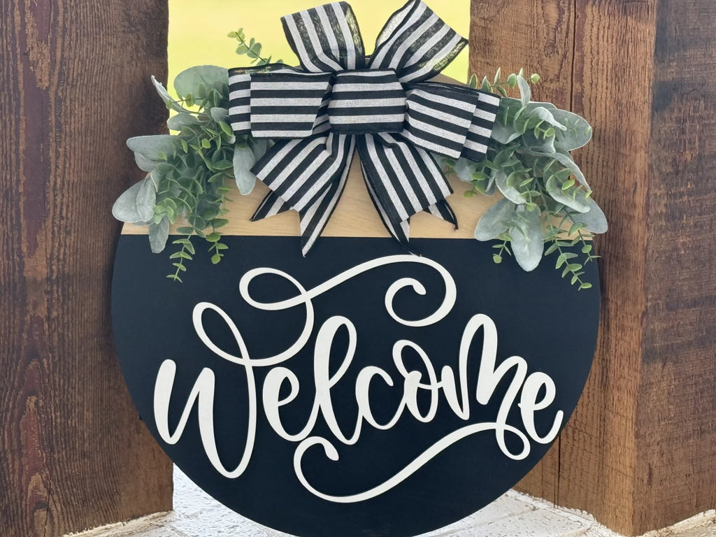 A round wooden sign with a black bottom half displaying “Welcome” in elegant white script. The top half is gold, decorated with green faux eucalyptus and a large black-and-white striped bow, leaning against a wooden background.