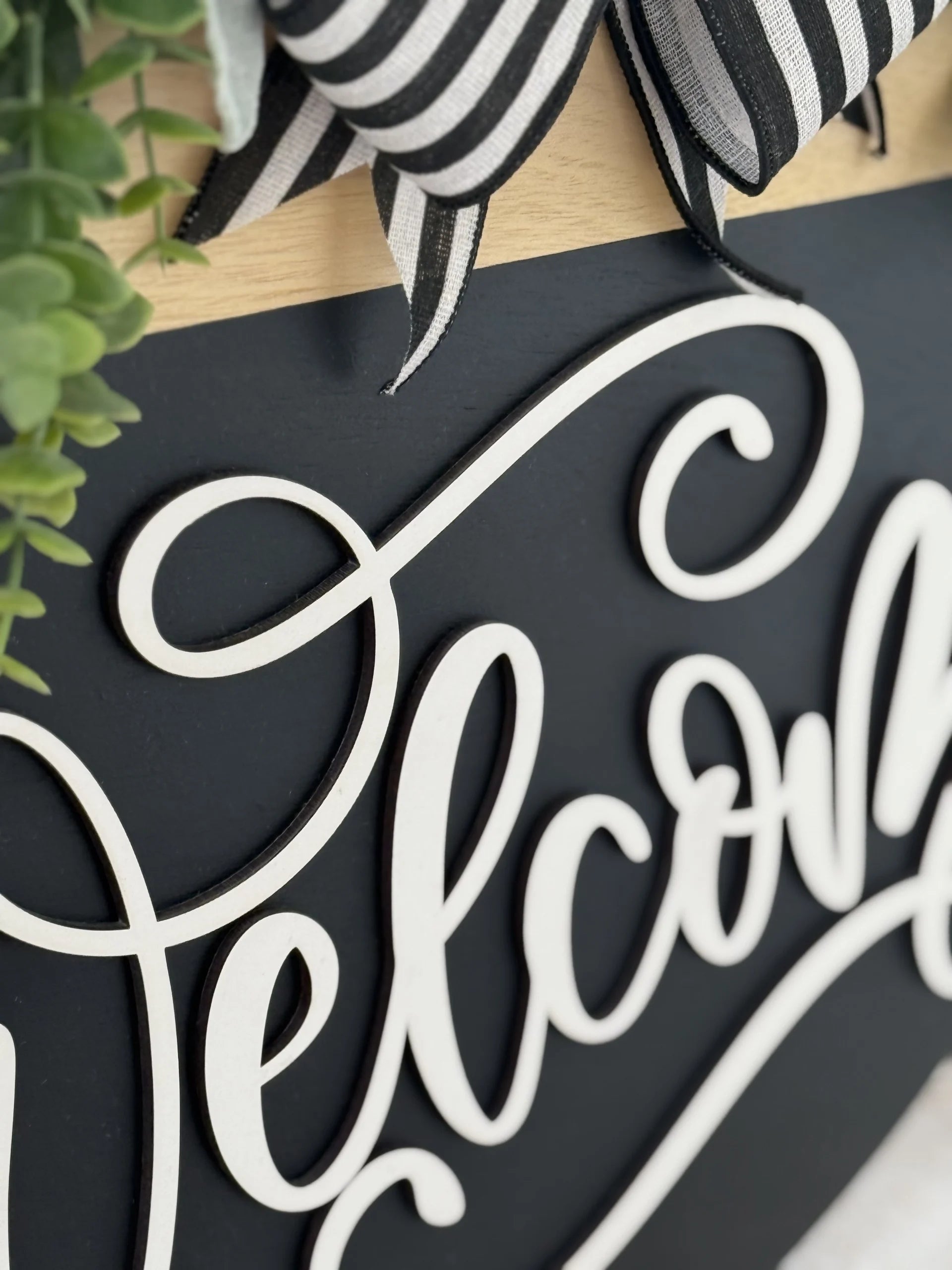 A close-up of a decorative welcome sign features elegant, white cursive lettering on a black background. Greenery and a black-and-white striped ribbon accent the sign at the top, adding a touch of natural and festive detail.