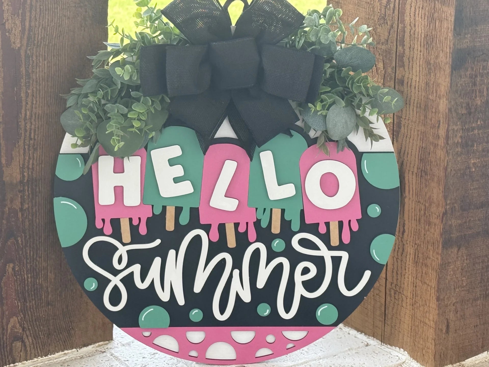 A round sign decorated with fake greenery and a black bow reads “HELLO Summer” in colorful letters. The word “HELLO” is on five pink popsicles, and “Summer” is written in white cursive. The sign has teal, pink, and white patterns and rests against wood.