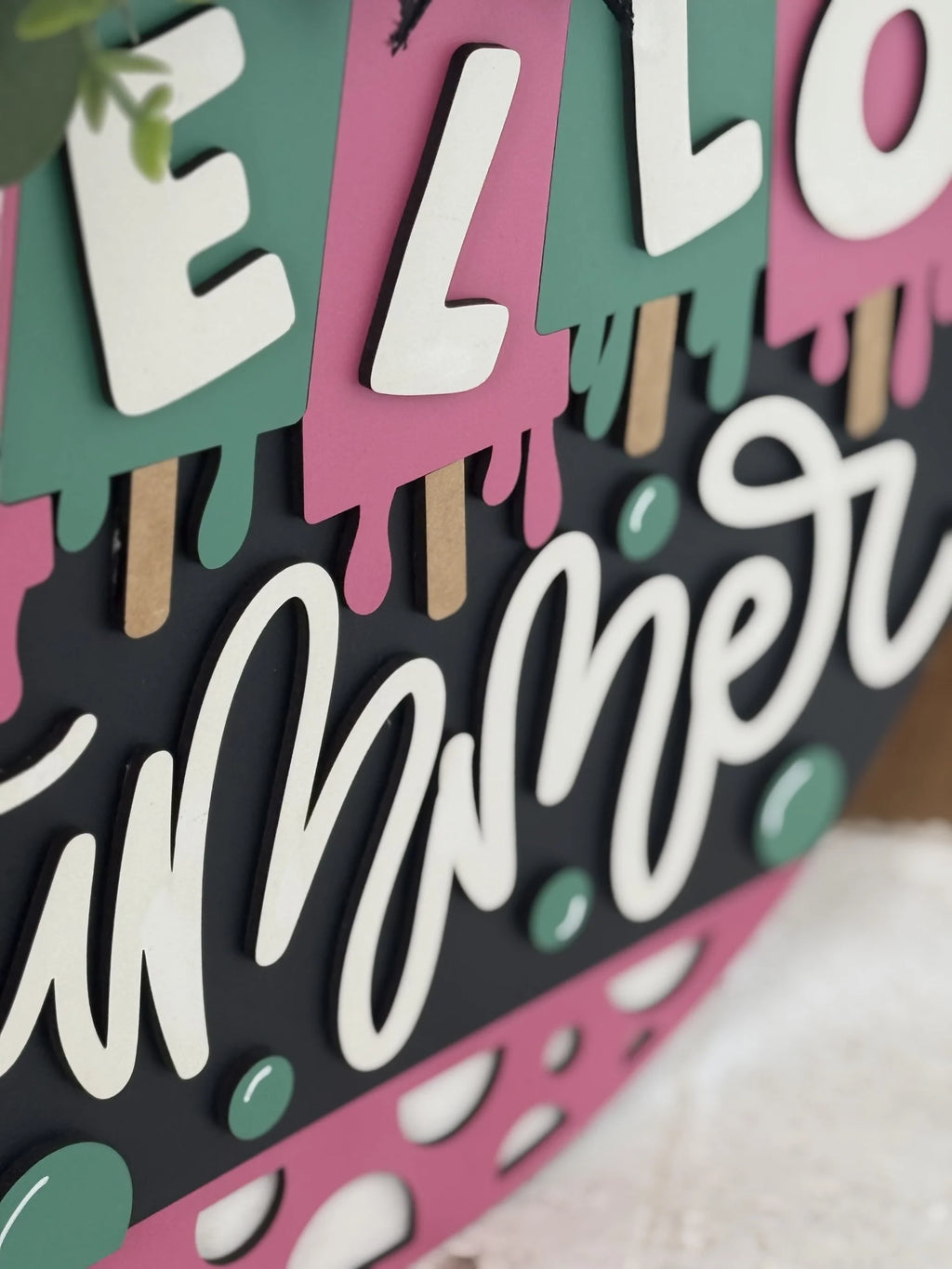 A close-up of a decorative sign features colorful popsicles in pink and green with white letters spelling HELLO and a cursive white summer below, accented with playful dots and popsicle sticks against a dark background.