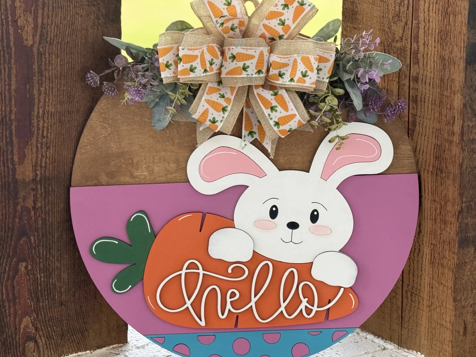 A wooden door sign features a smiling white bunny holding an orange carrot with hello written on it. The bunny is in front of a purple and turquoise background with pink dots. At the top, a bow decorated with carrots and greenery accents is attached.