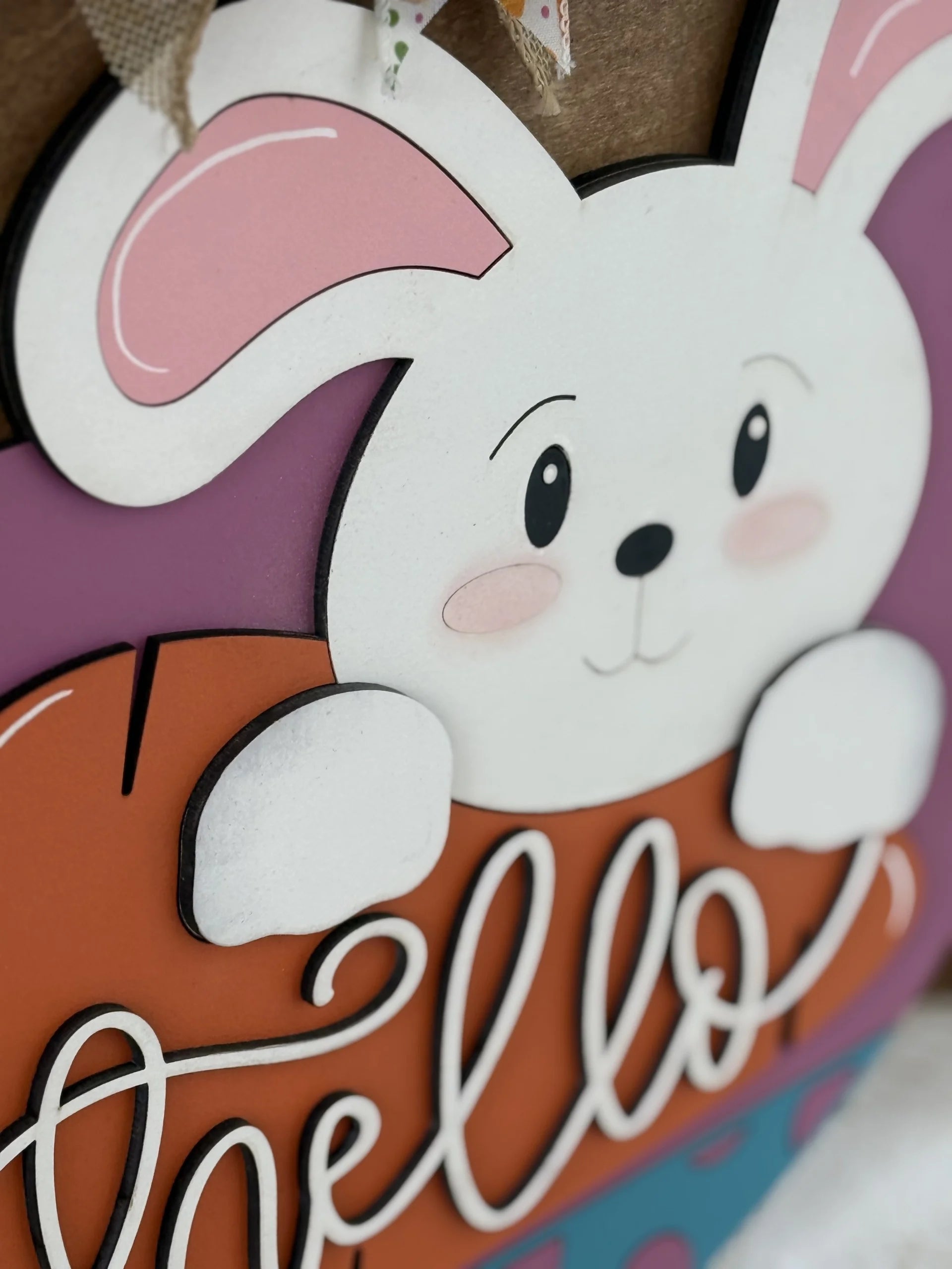 A close-up of a decorative sign shows a white cartoon bunny with pink ears and rosy cheeks, holding an orange shape with the word hello written in cursive. The background includes purple and blue accents and a burlap ribbon at the top.