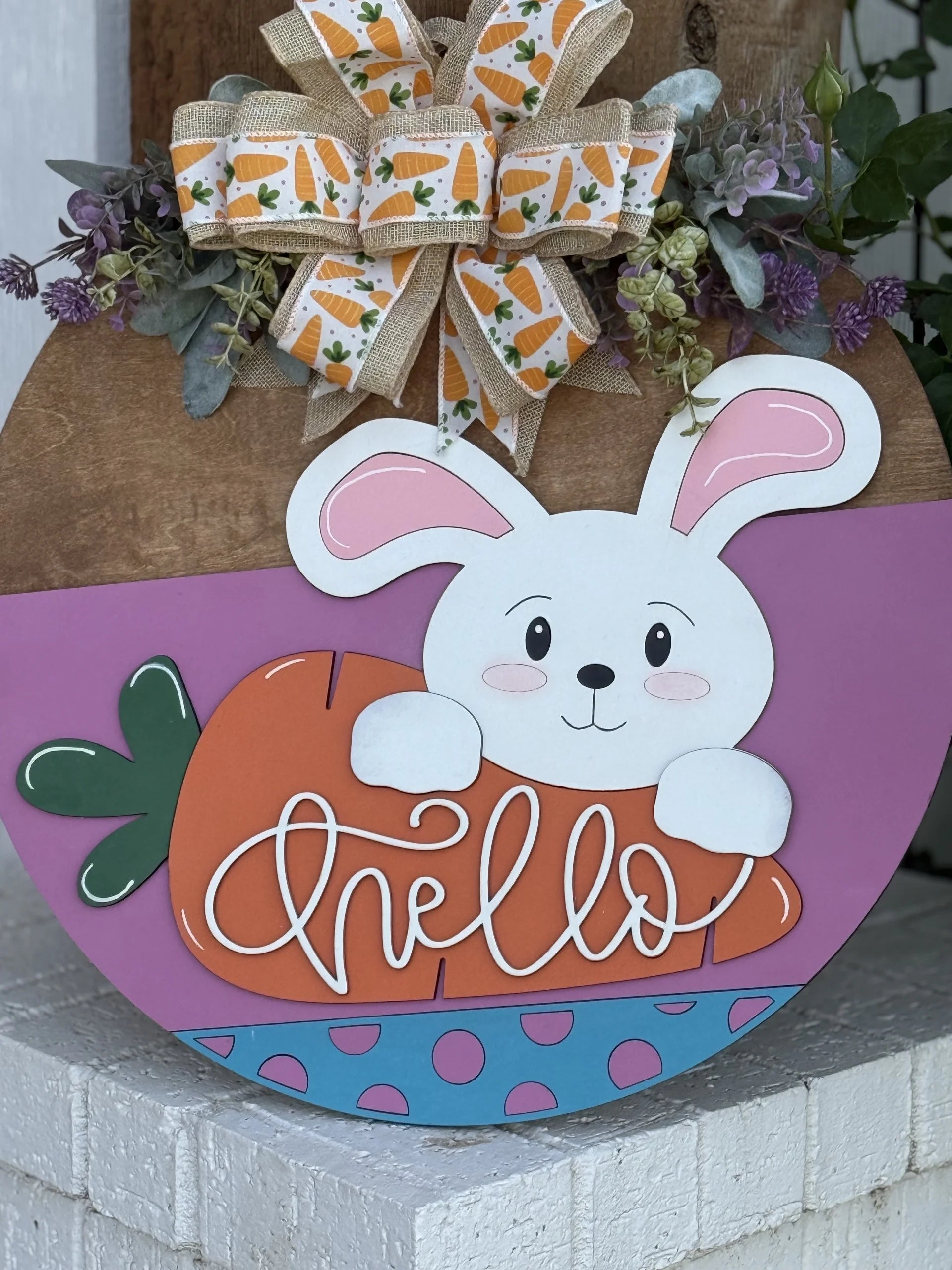 A round Easter-themed door sign features a cute white bunny holding an orange carrot with “hello” written on it in cursive. The sign has purple and blue sections, and is topped with a bow decorated with carrots and greenery.