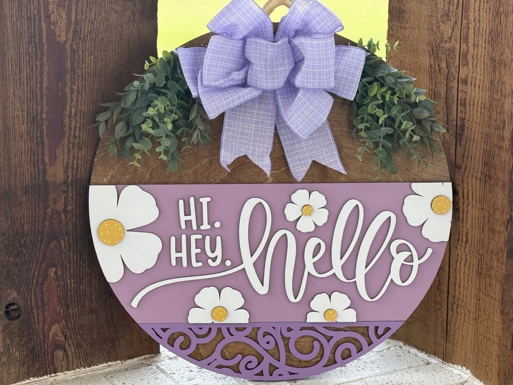 A round wooden door sign features a large purple plaid bow, artificial greenery, and white flowers with yellow centers. The sign reads HI. HEY. hello in white decorative letters on a lavender background with ornate cutout patterns at the bottom.