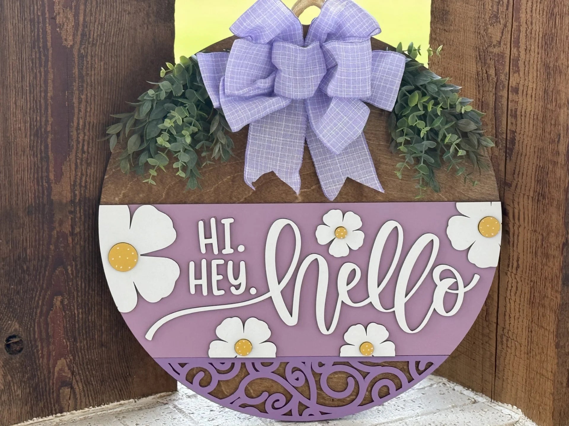 A round wooden door sign features a large purple plaid bow, artificial greenery, and white flowers with yellow centers. The sign reads HI. HEY. hello in white decorative letters on a lavender background with ornate cutout patterns at the bottom.