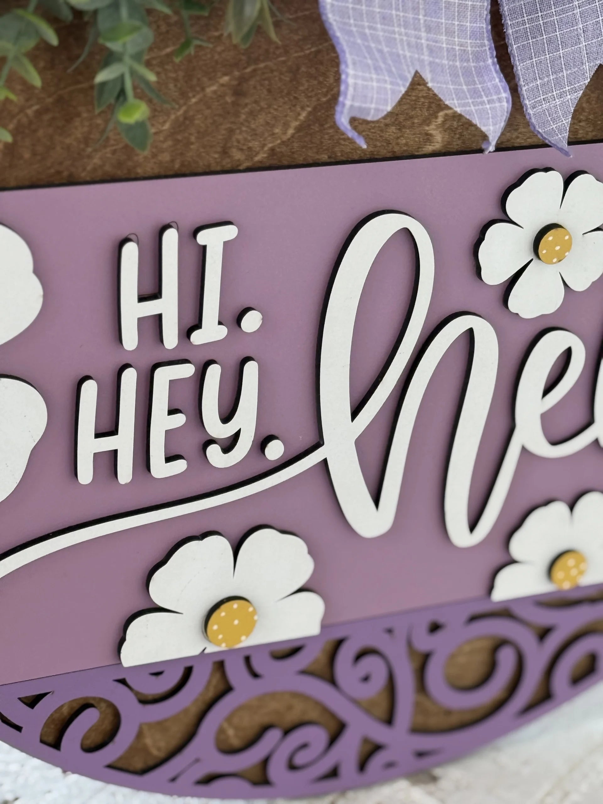 Close-up of a decorative sign with a lavender background and white, raised text reading HI. HEY. HE with floral designs. Part of a purple plaid ribbon and green leaves are visible at the top, with white flowers featuring yellow centers as accents.