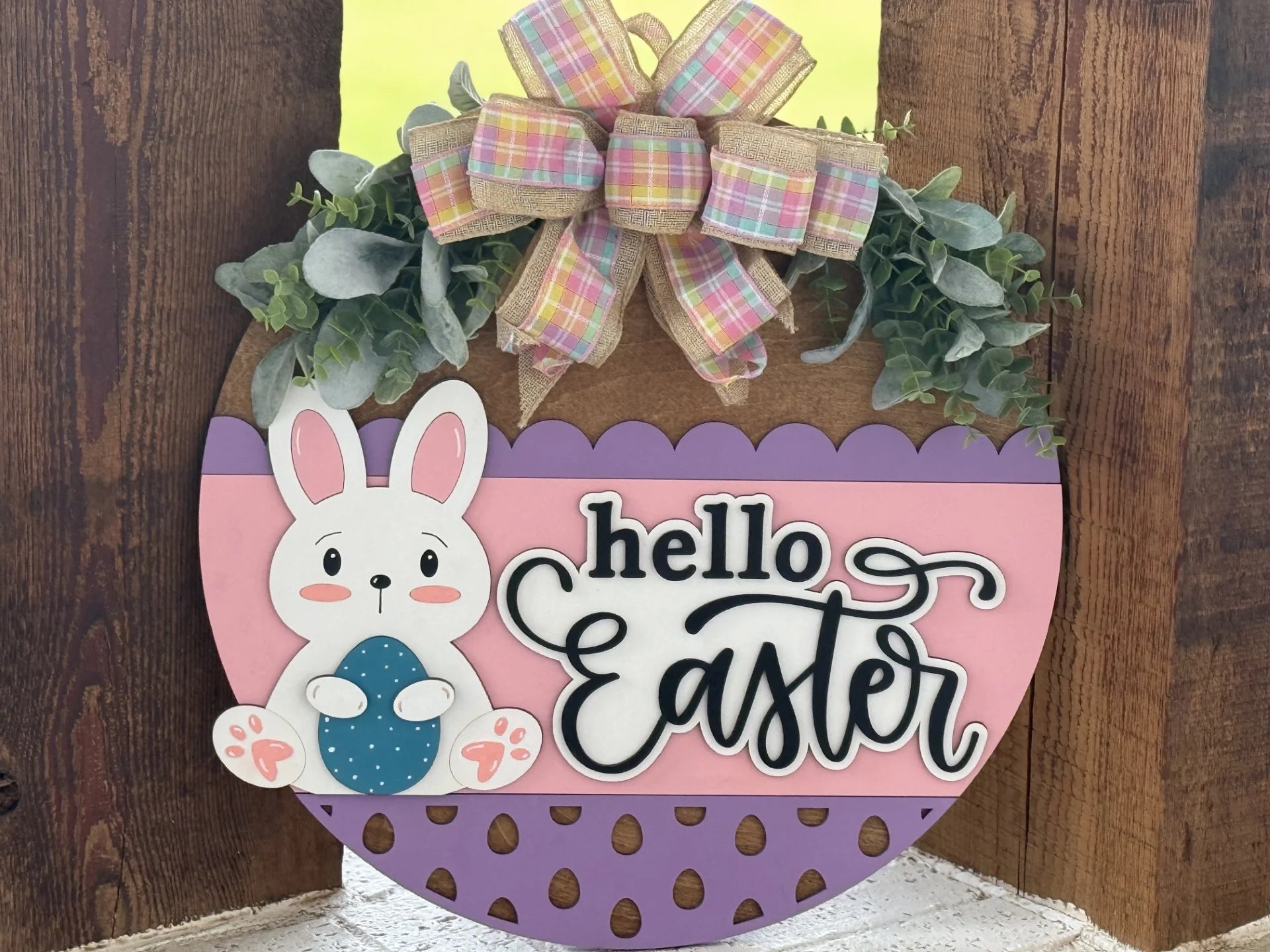 A wooden Easter decoration shaped like an egg features a cute white bunny holding a blue egg, pastel colors, green leaves, a plaid bow, and the words hello Easter in black script on a pink and purple background. It leans against a wooden wall.