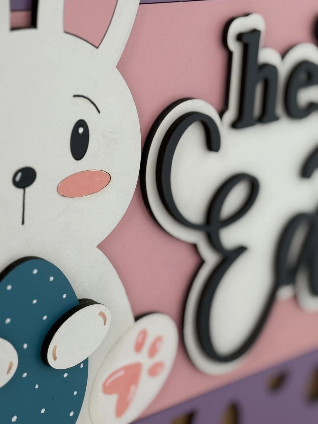 Close-up of a cute white cartoon bunny holding a blue decorated egg. The bunny has pink ears, cheeks, and paw, with black eyes and nose. Part of raised black-and-white text and a pink background are visible, suggesting a decorative Easter sign.