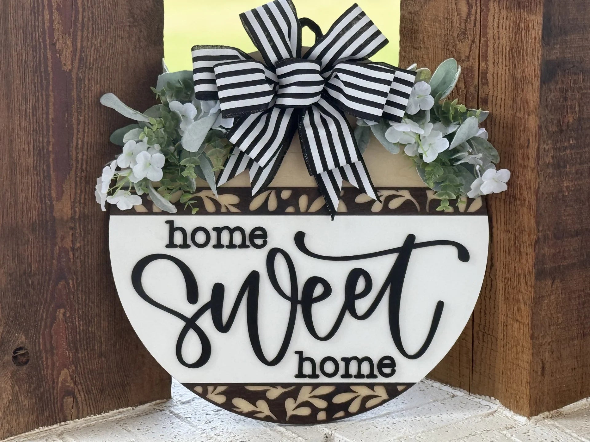 A round decorative sign features the words home sweet home in black script and print. The top is adorned with white flowers, greenery, and a large black-and-white striped bow. The background is wood with a white center and floral border accents.