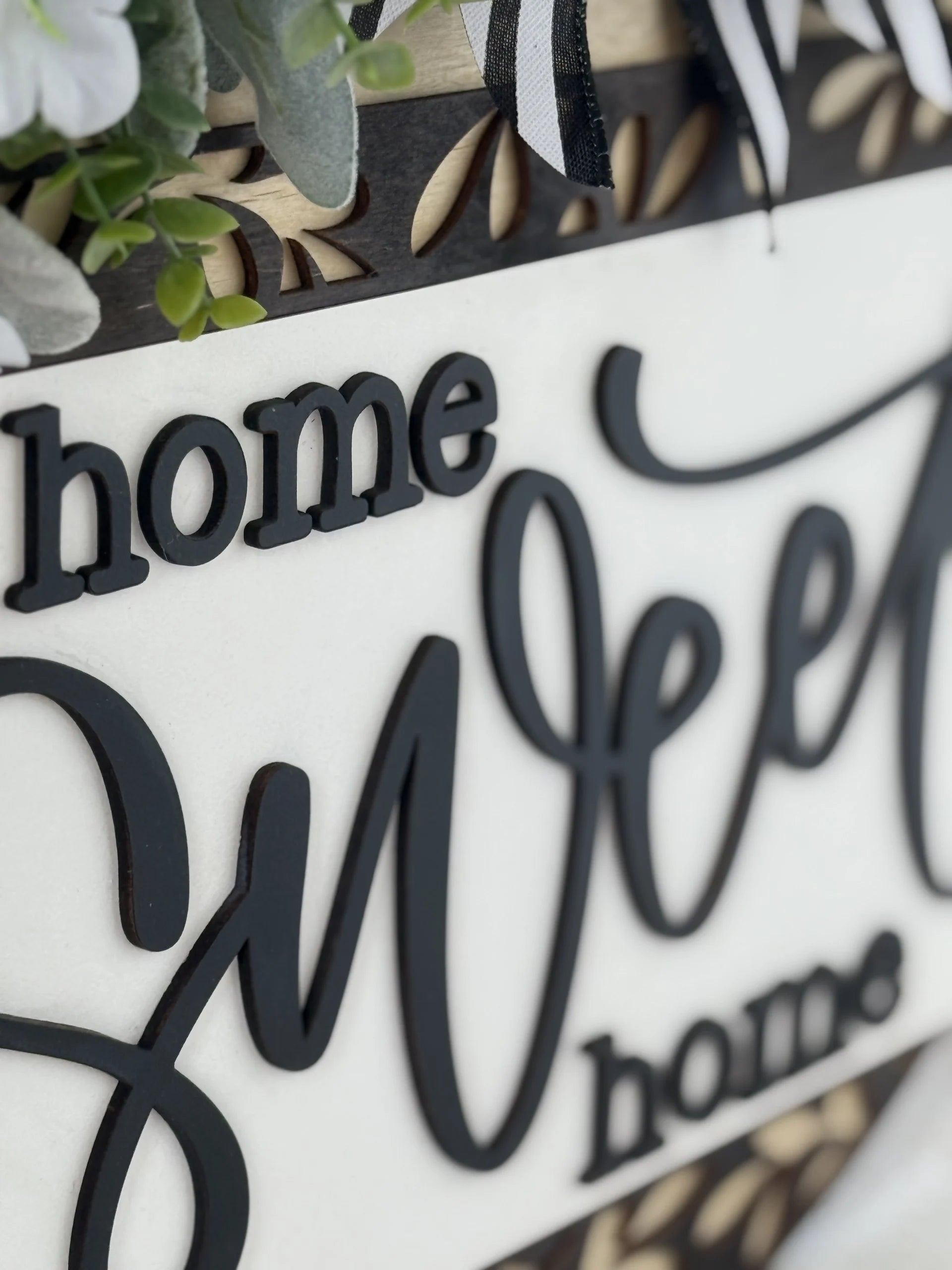 Close-up of a decorative sign with raised, black script letters spelling “home sweet home” on a white background. Greenery, white flowers, and a black-and-white striped ribbon accent the top edge, adding a welcoming and cozy touch.