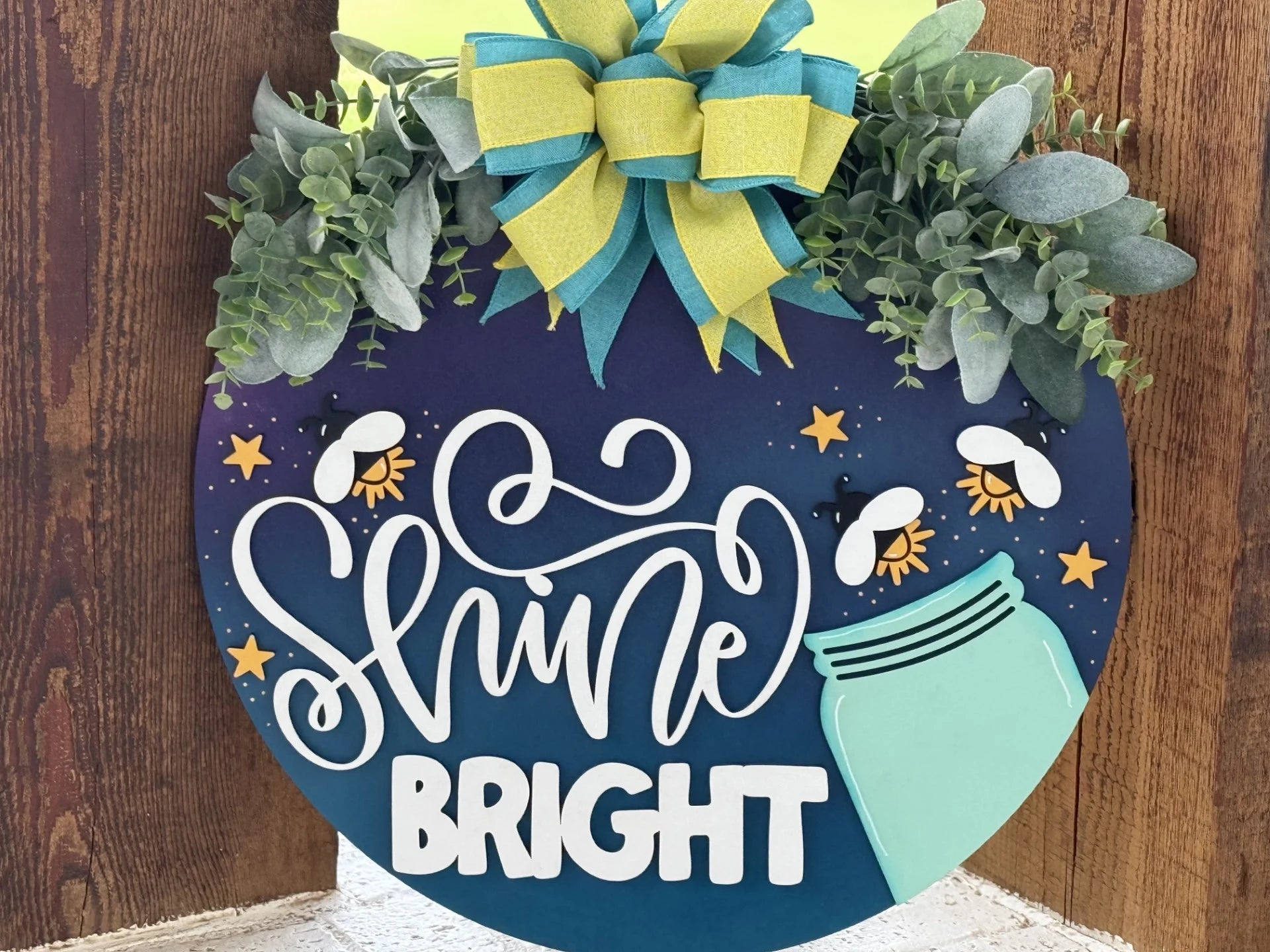 A round sign with greenery and a large blue-and-yellow bow on top reads “Shine Bright” in white script. The background is blue with yellow stars, fireflies, and a mint green jar, evoking a cheerful, summer night theme. The sign leans against a wood wall.