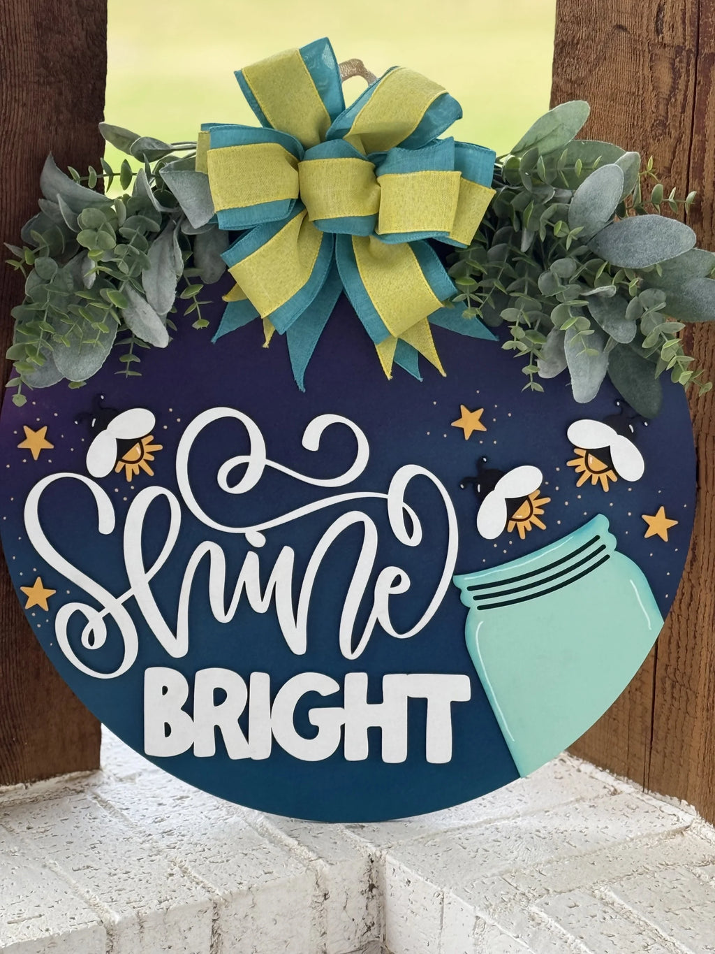 A round navy-blue sign decorated with faux greenery and a blue-green-yellow bow reads “Shine Bright” in bold white script. Yellow stars, fireflies, and a turquoise mason jar design accent the sign, which hangs on a wooden post.